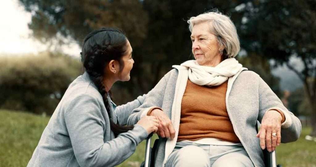 Caregiver chatting with a senior in a wheelchair outdoors.